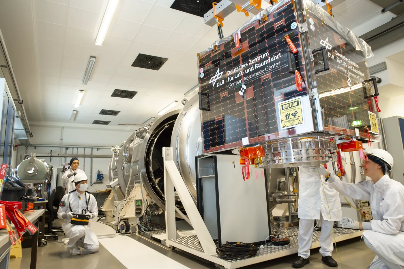 A drum-shaped satellite covered in solar cells standing on a frame in a cleanroom beside a thermal vacuum chamber, with three technicians in white coveralls working around it